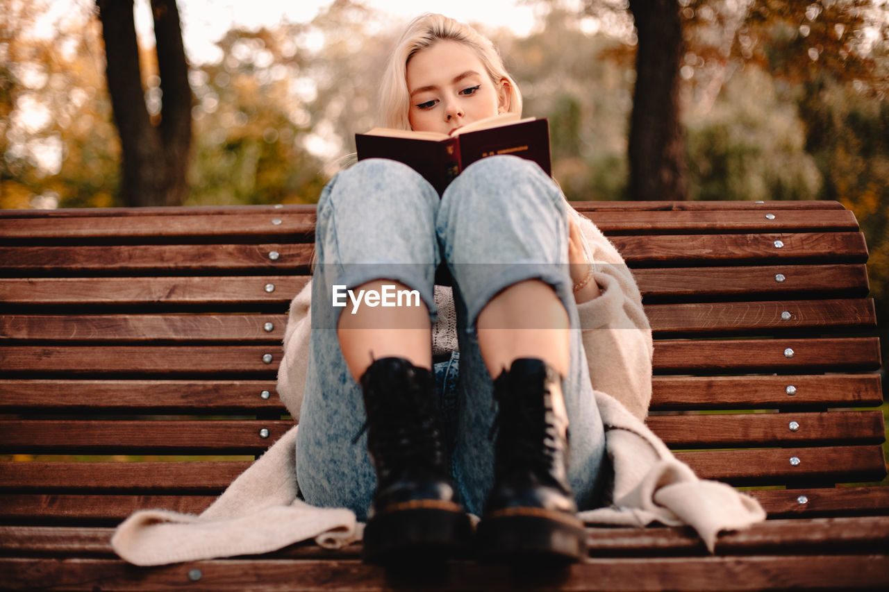 Teenage girl reading book while sitting on bench in park during autumn