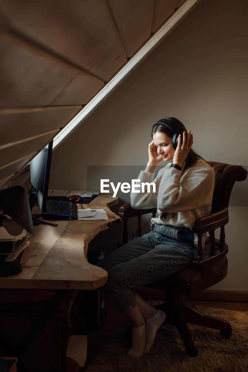 Young woman listening to music in big headphones near computer at home. creating play list, enjoying