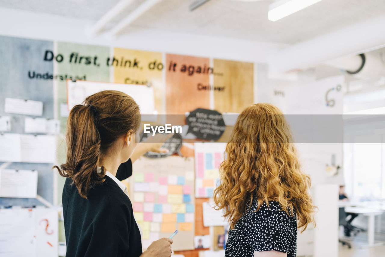 Confident female colleagues discussing over strategy on wall at creative office