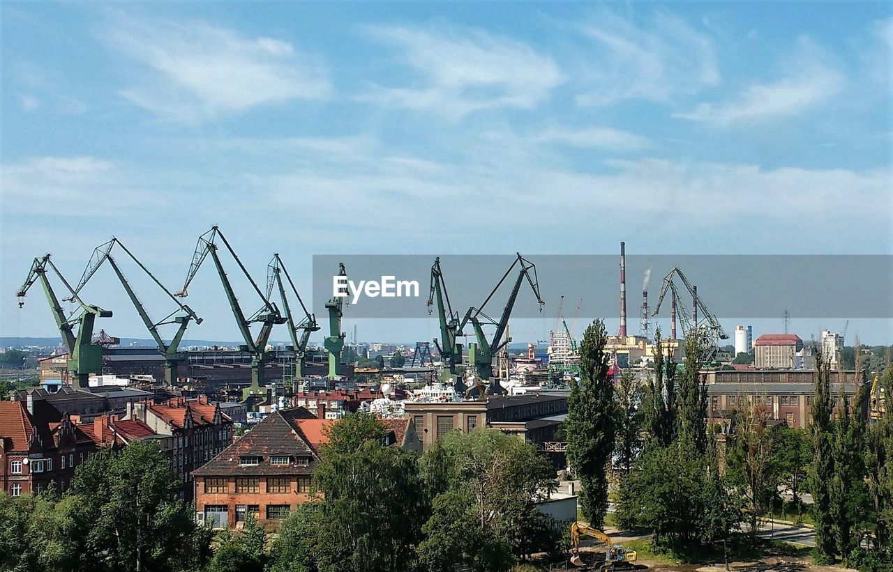 Buildings by cranes at harbor against sky