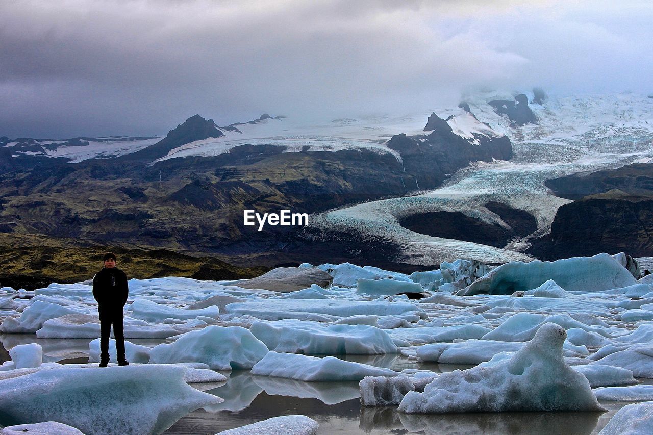 Full length man standing on ice by snow covered mountains against cloudy sky