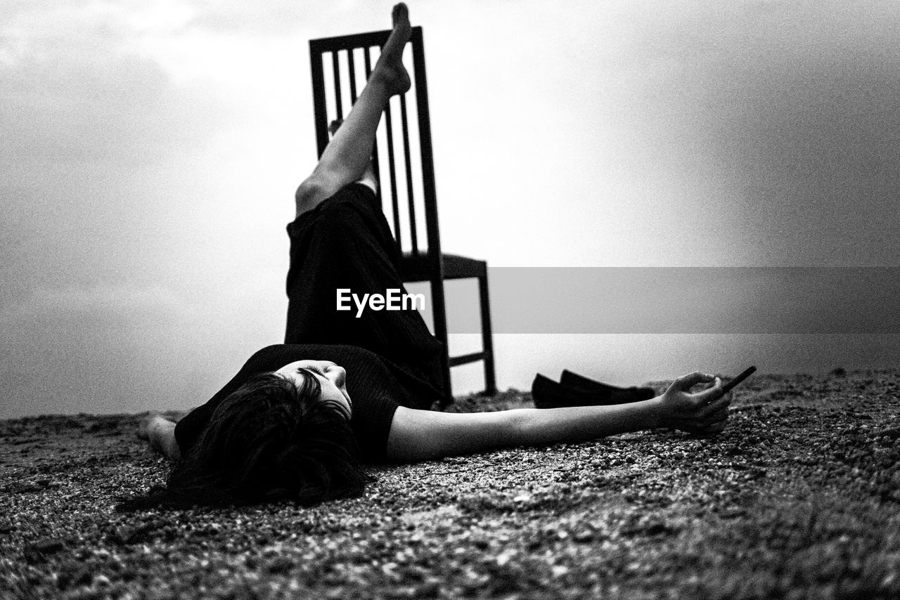 Woman lying on shore by chair against sky