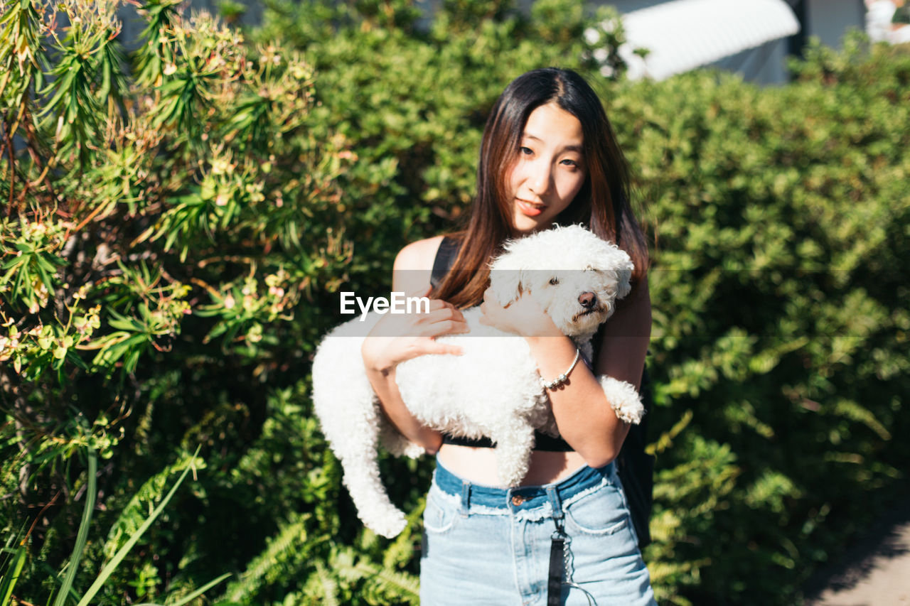 Portrait of smiling young woman carrying white dog against plants during sunny day