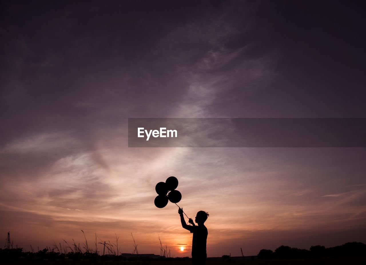 Silhouette boy holding balloons against sky during sunset