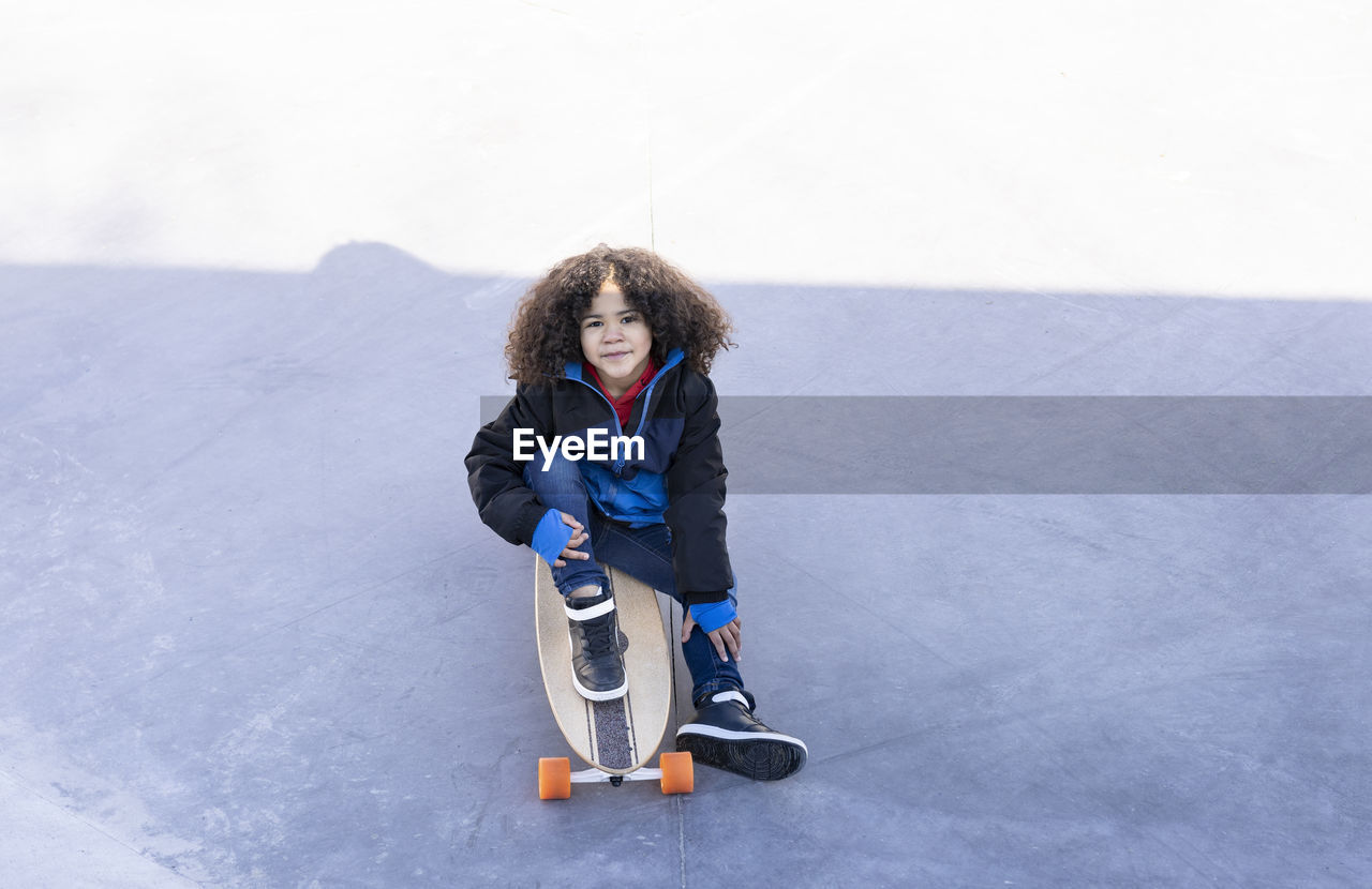 Ethnic child with curly hair sitting on ramp with longboard in skate park and looking at camera