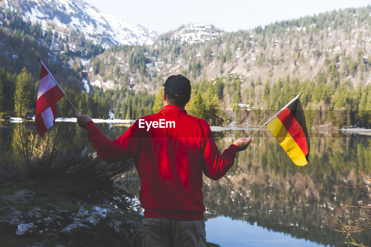 Rear view of mature man holding national flags while standing by lake against mountain