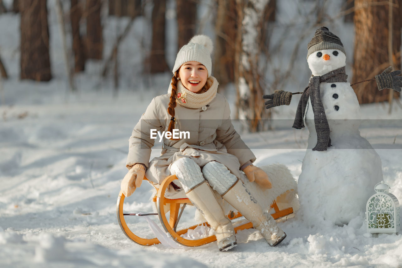 Portrait of cute girl sitting by snowman in forest
