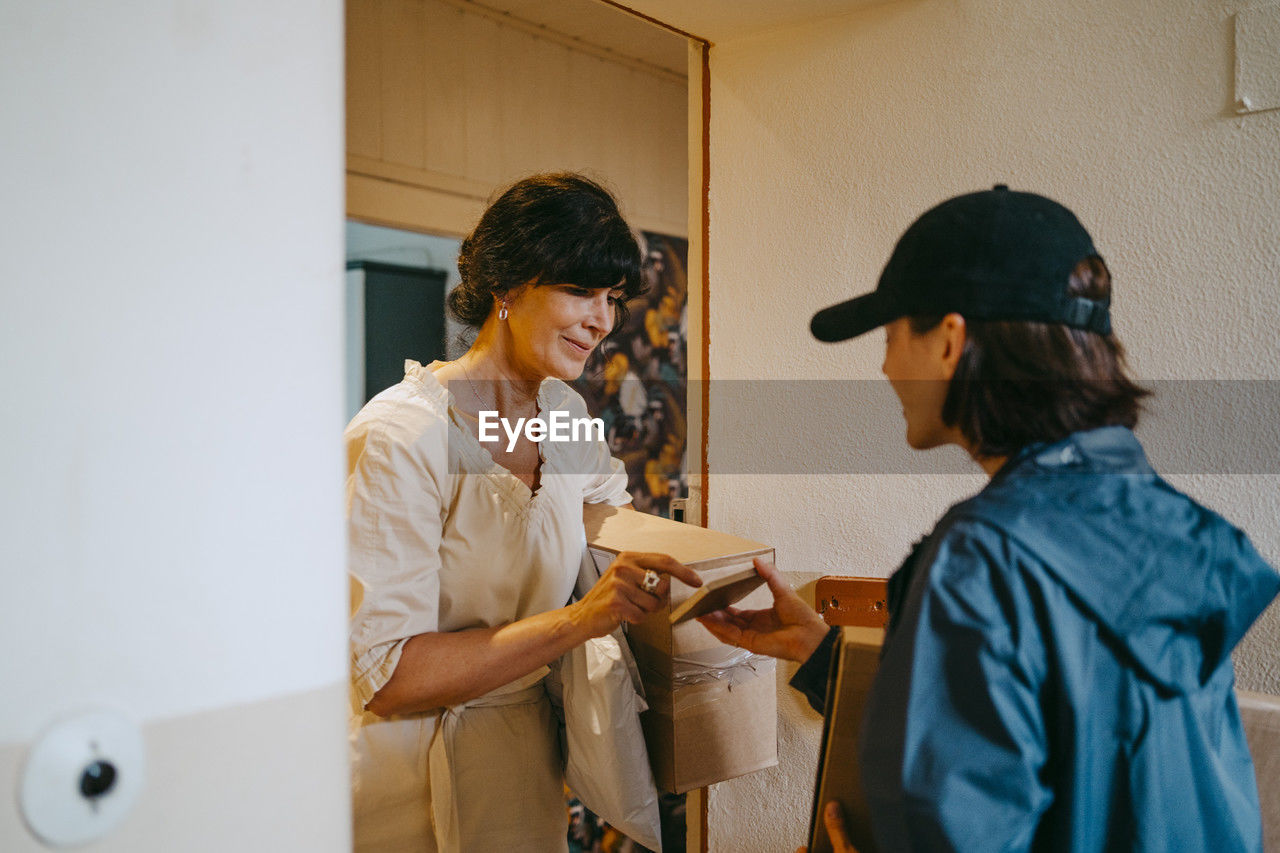 Mature woman doing digital signature while receiving parcel from delivery person at house
