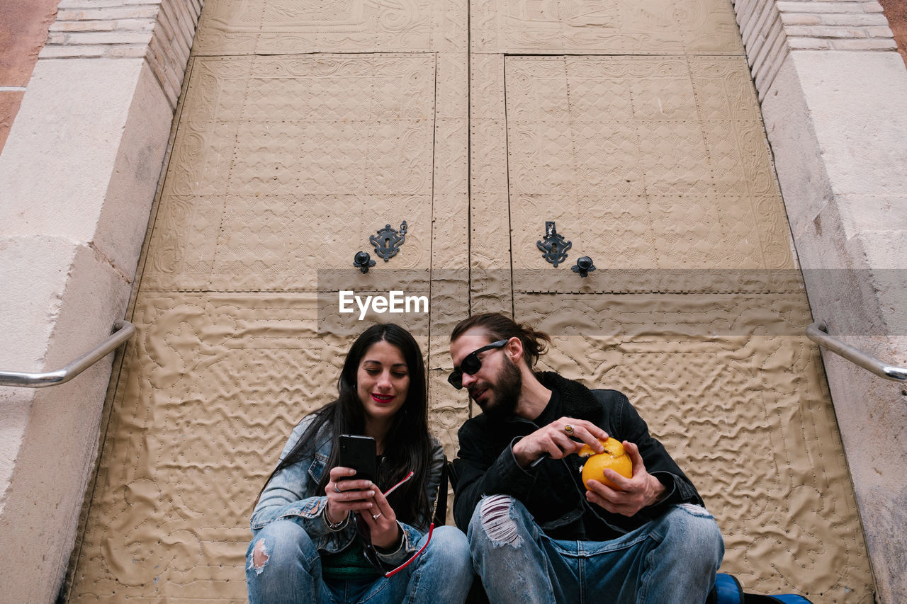 Man and woman staring at the phone eating fruit sitting in the street