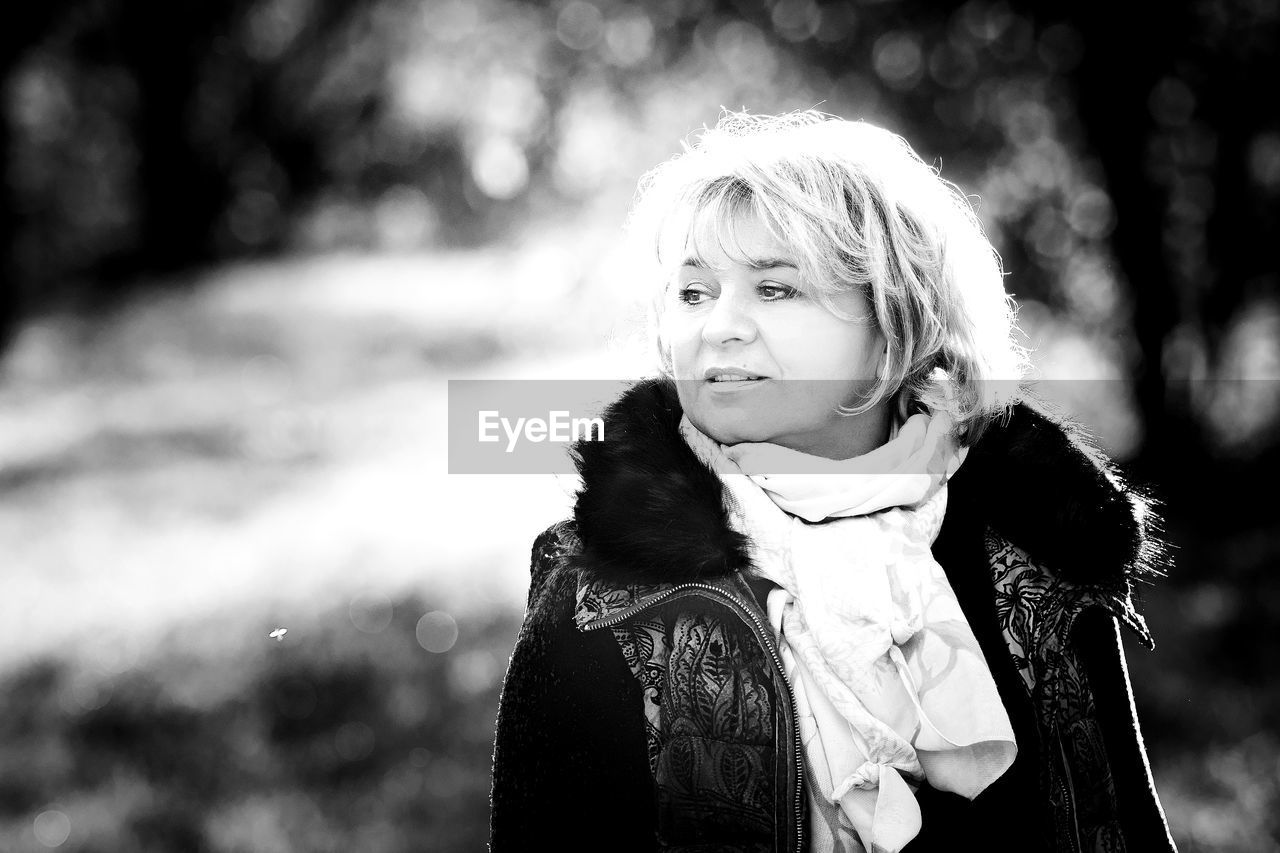 PORTRAIT OF SMILING WOMAN STANDING AGAINST BLURRED BACKGROUND