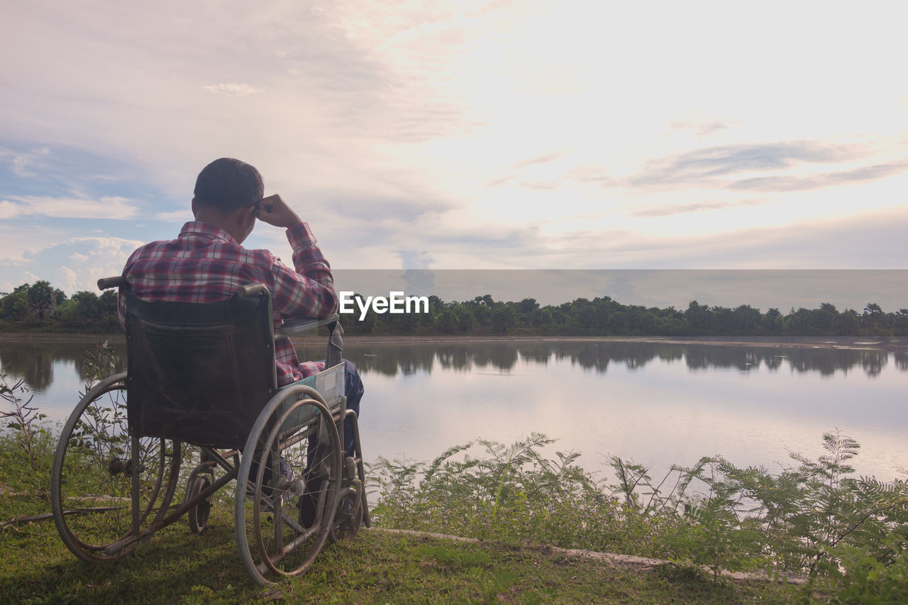 REAR VIEW OF MAN SITTING IN LAKE AGAINST SKY