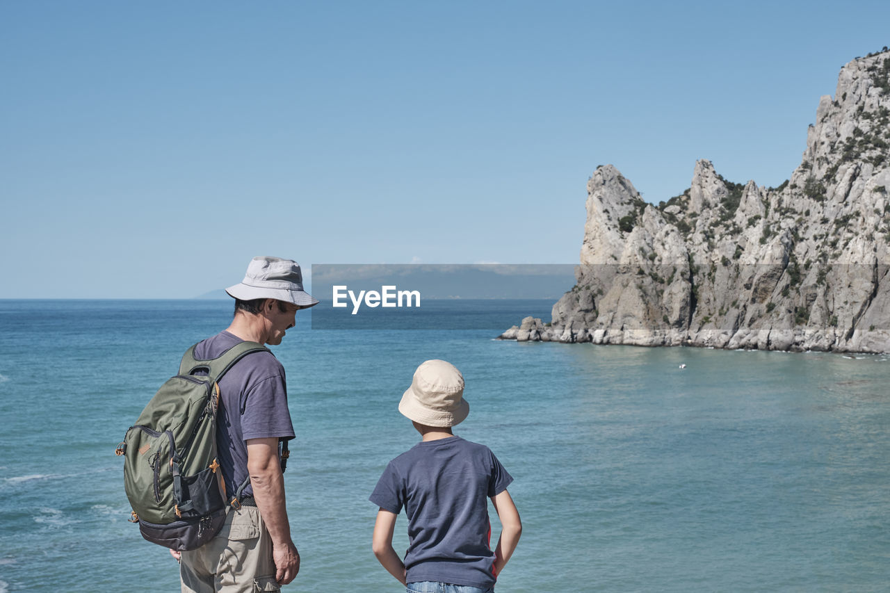 A boy and his grandfather standing on the shore of the bay, talking, looking at the rock. 