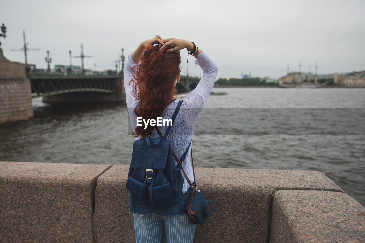 Back view of unrecognizable red haired female tourist with backpack standing near stone fence against river and enjoying wind while resting during sightseeing in cloudy day in saint petersburg in russia