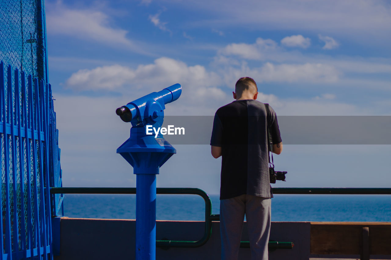 Rear view of man standing by coin-operated binoculars at railing against sea at observation point