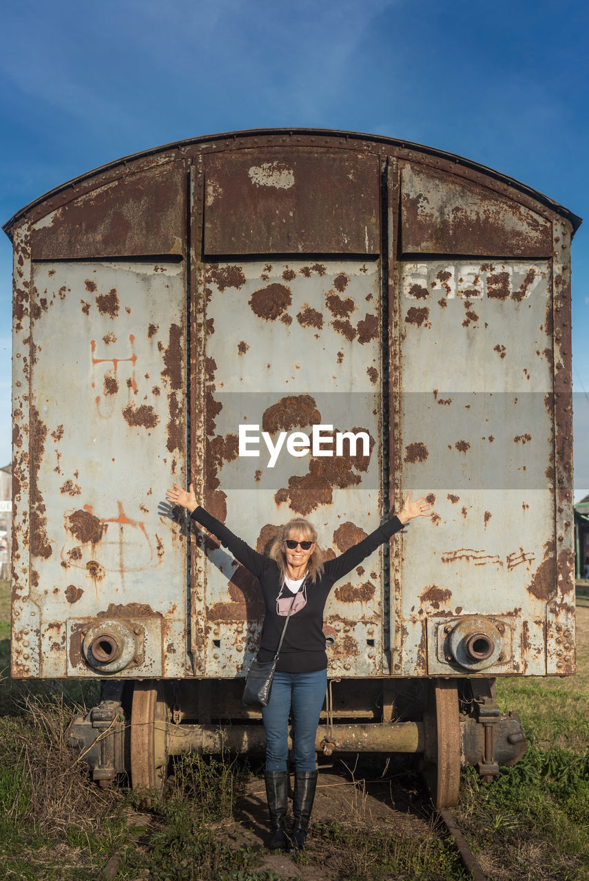 Adult woman wearing sunglasses smiling with her arms raised and abandoned train carriage