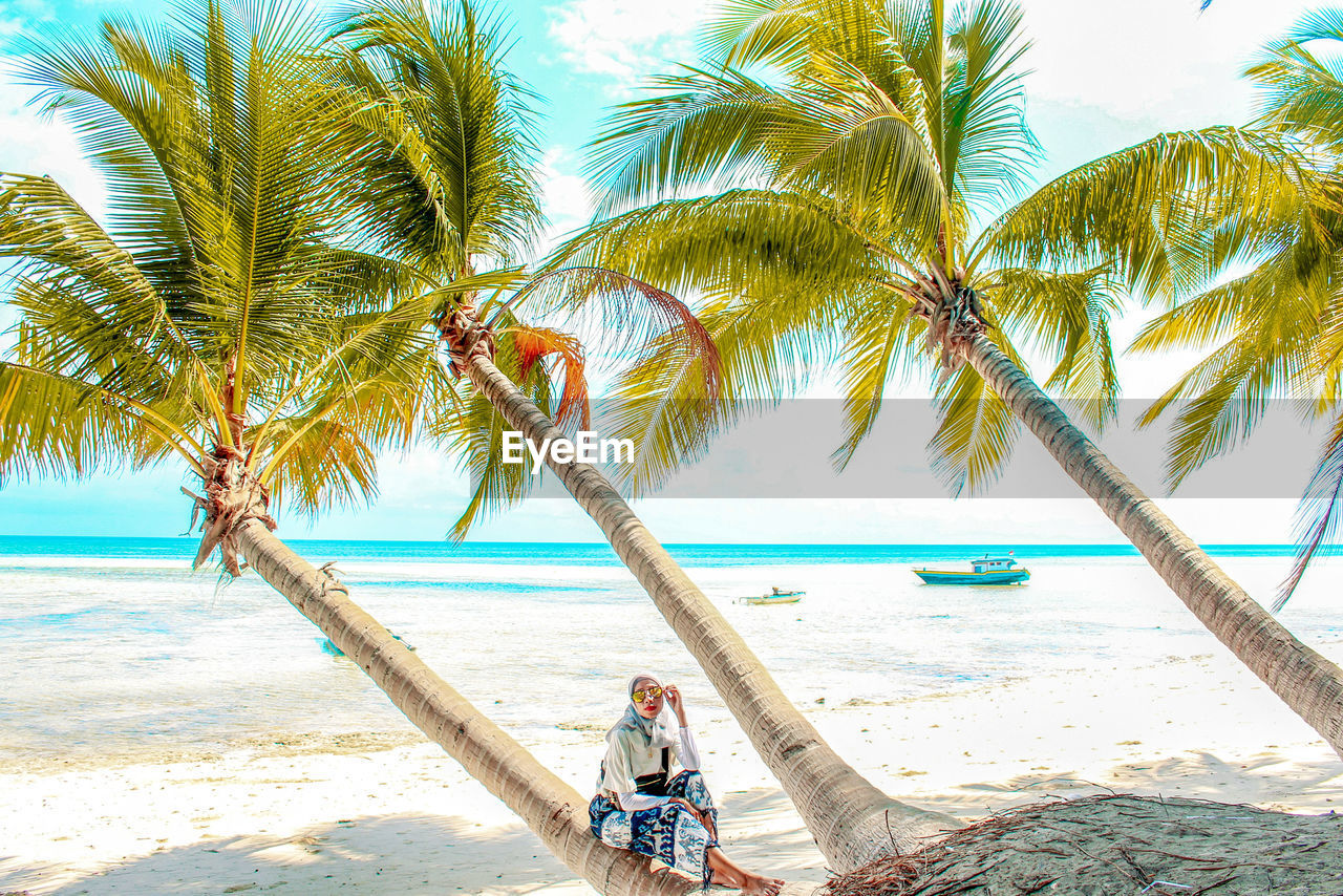 Woman sitting on palm trees at beach against sky