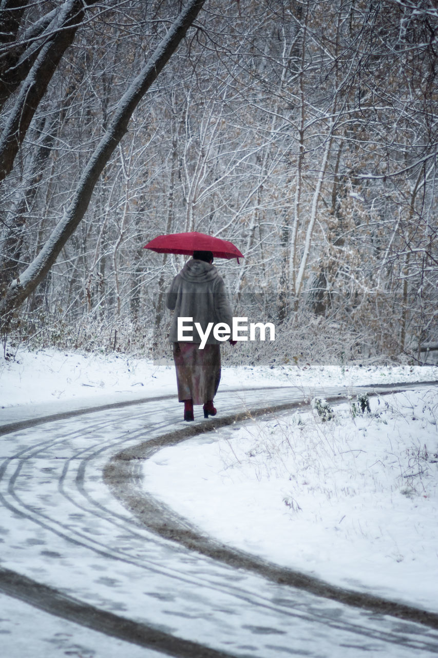 Woman in a gray fur coat with a red umbrella walks through a snow-covered park.