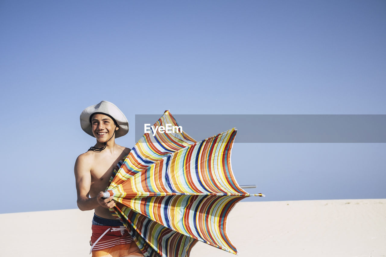 Shirtless teenage boy holding umbrella at beach against clear blue sky