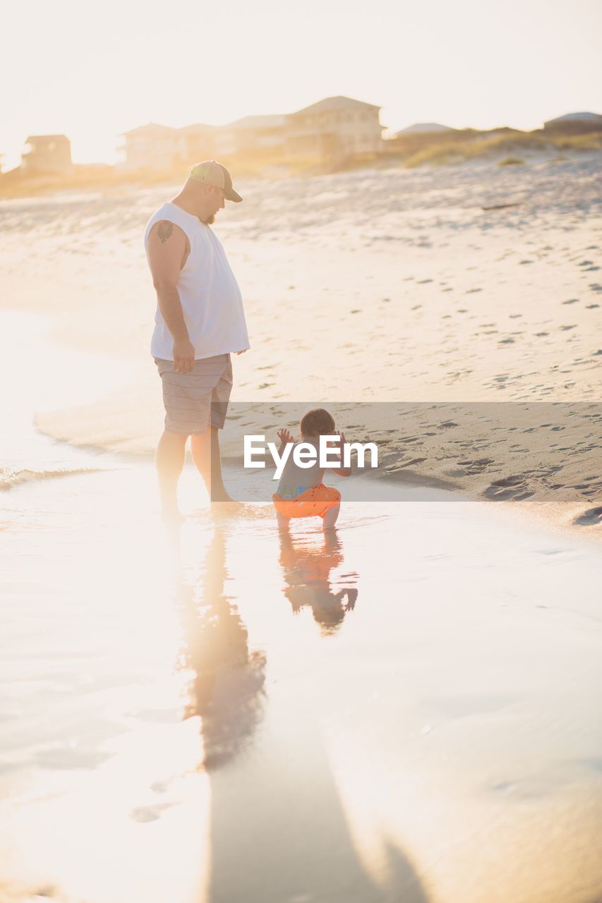 Father and son standing at beach against sky during sunset