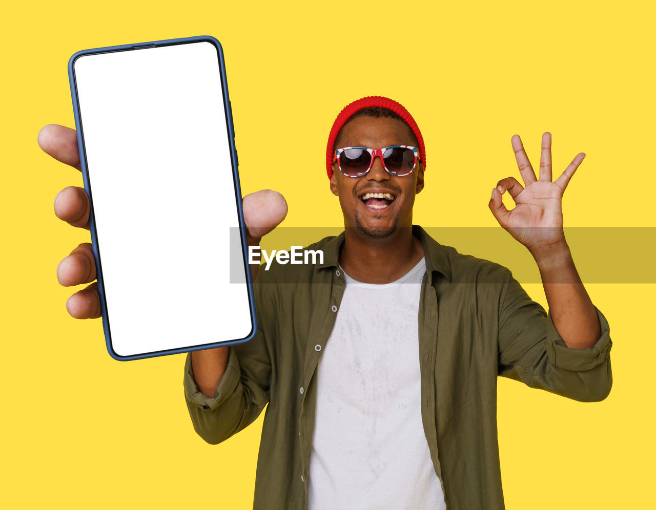 smiling, one person, glasses, fashion, happiness, technology, adult, communication, sunglasses, yellow, finger, wireless technology, portrait, men, cheerful, thumbs up, emotion, front view, young adult, gesturing, studio shot, showing, fun, sign language, positive emotion, casual clothing, standing, cartoon, hand sign, looking at camera, waist up, cool attitude, cut out, holding, colored background, person, copy space, funky, enjoyment, indoors, youth culture, portable information device, clothing, smile, smartphone, teeth, arts culture and entertainment