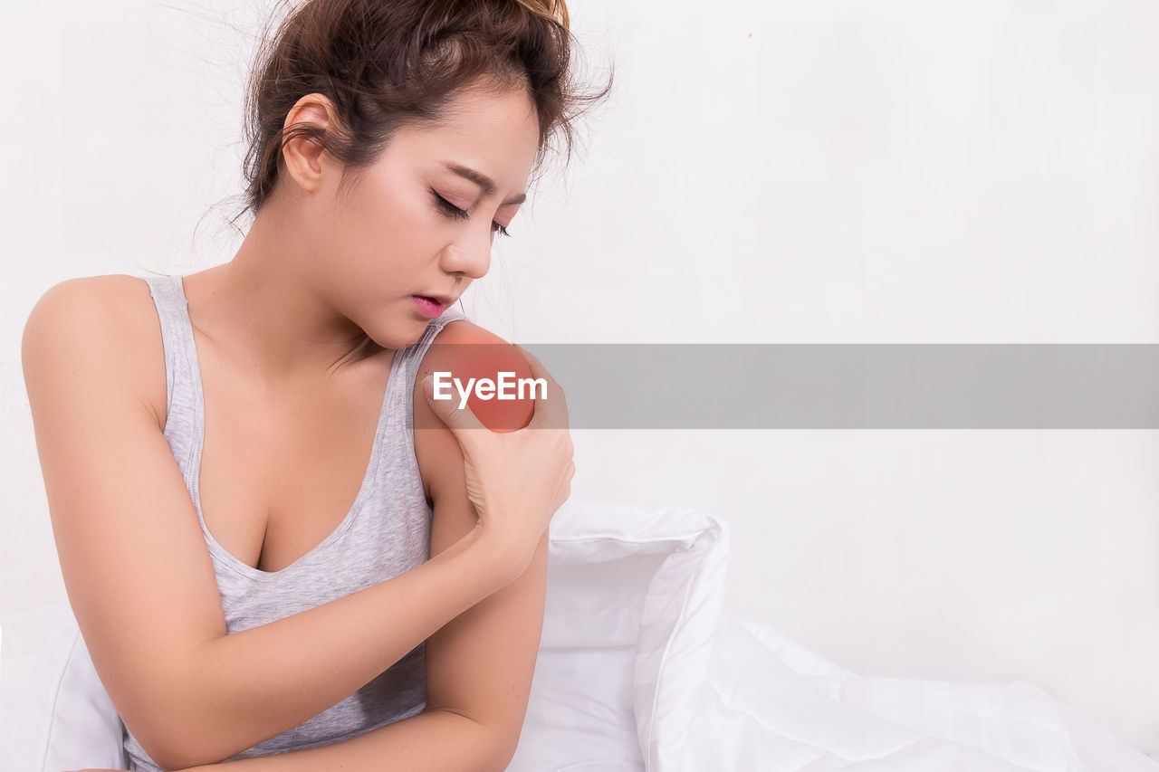 Young woman holding shoulder while sitting on bed at home