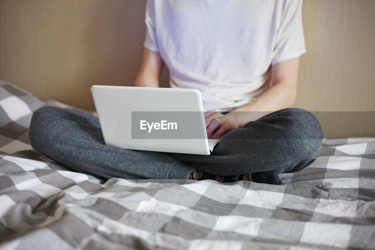 MIDSECTION OF WOMAN USING MOBILE PHONE WHILE SITTING ON BED