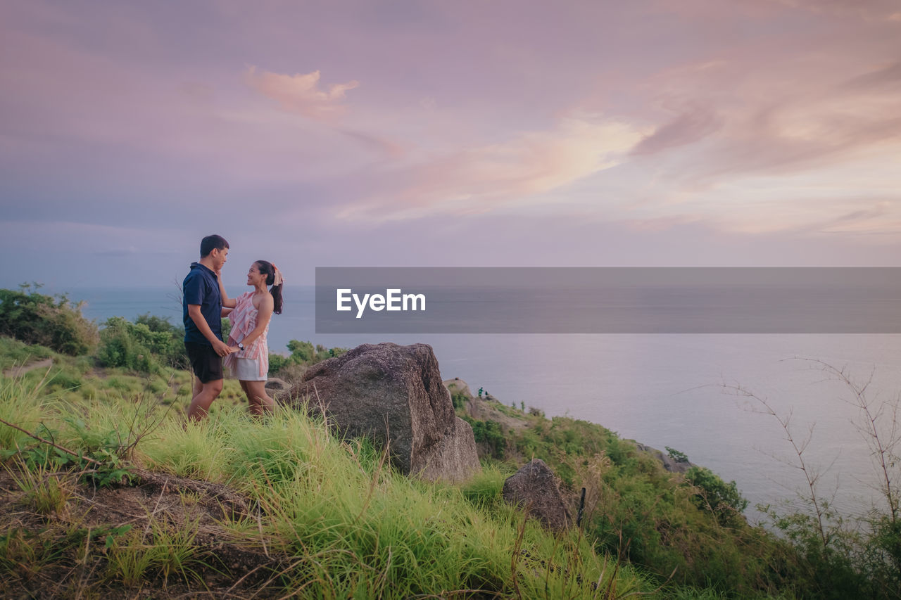 Rear view of man standing on rock by sea against sky during sunset