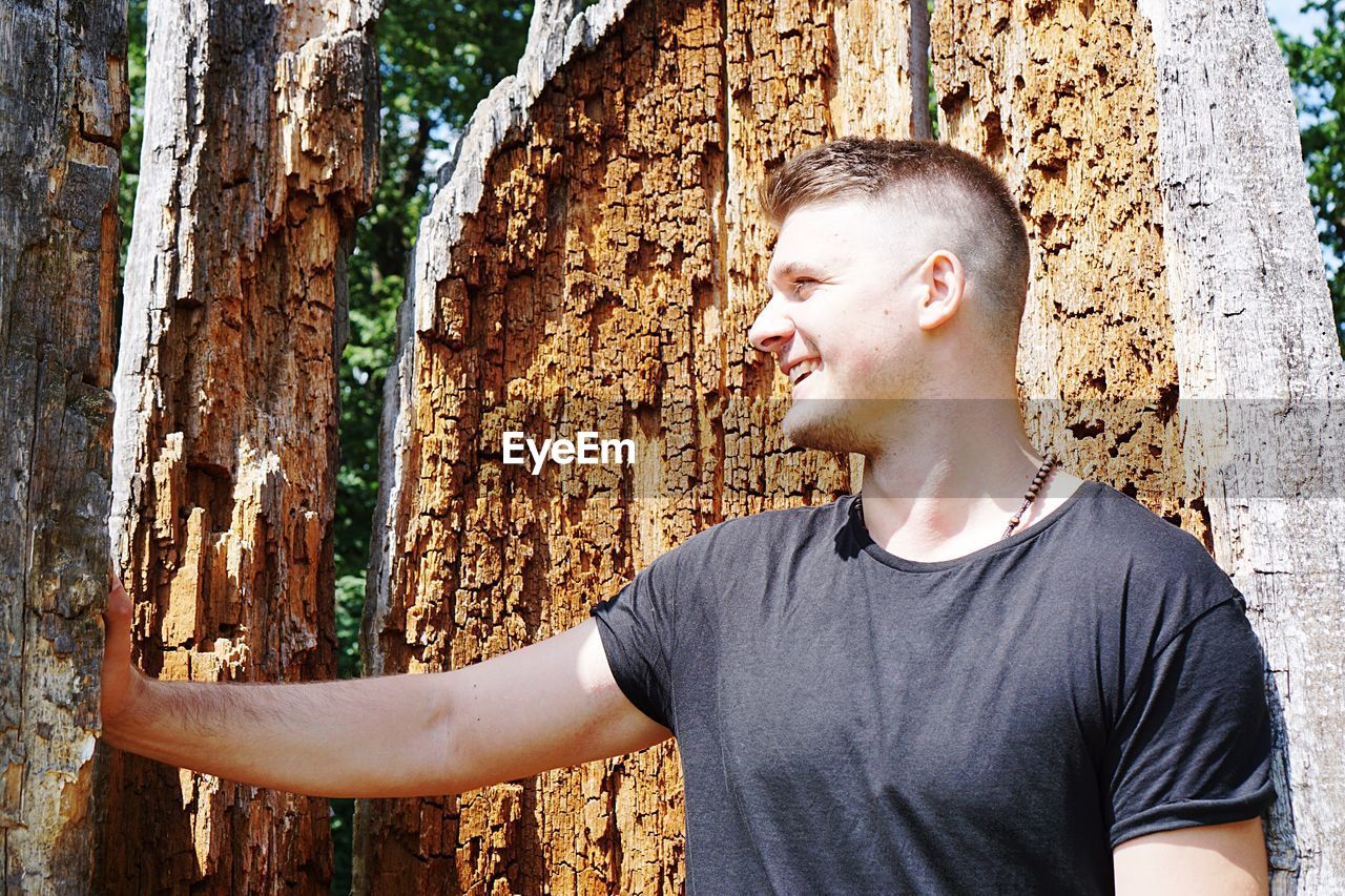 Young man looking away while standing against tree trunk