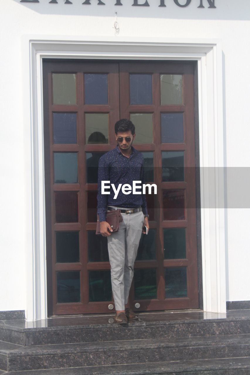 FULL LENGTH PORTRAIT OF YOUNG MAN STANDING AGAINST BUILDING