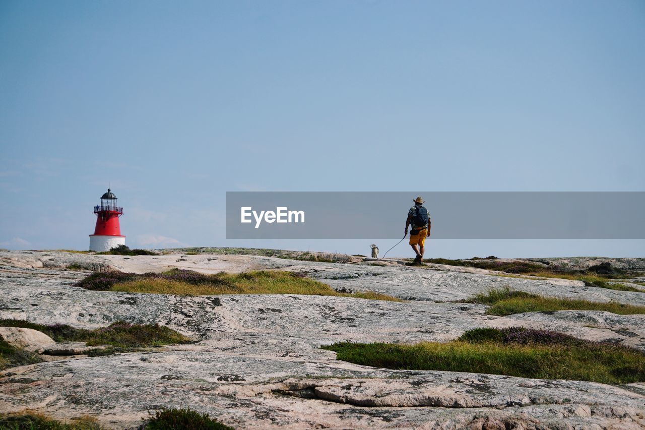 Rear view of man with dog walking towards lighthouse on mountain