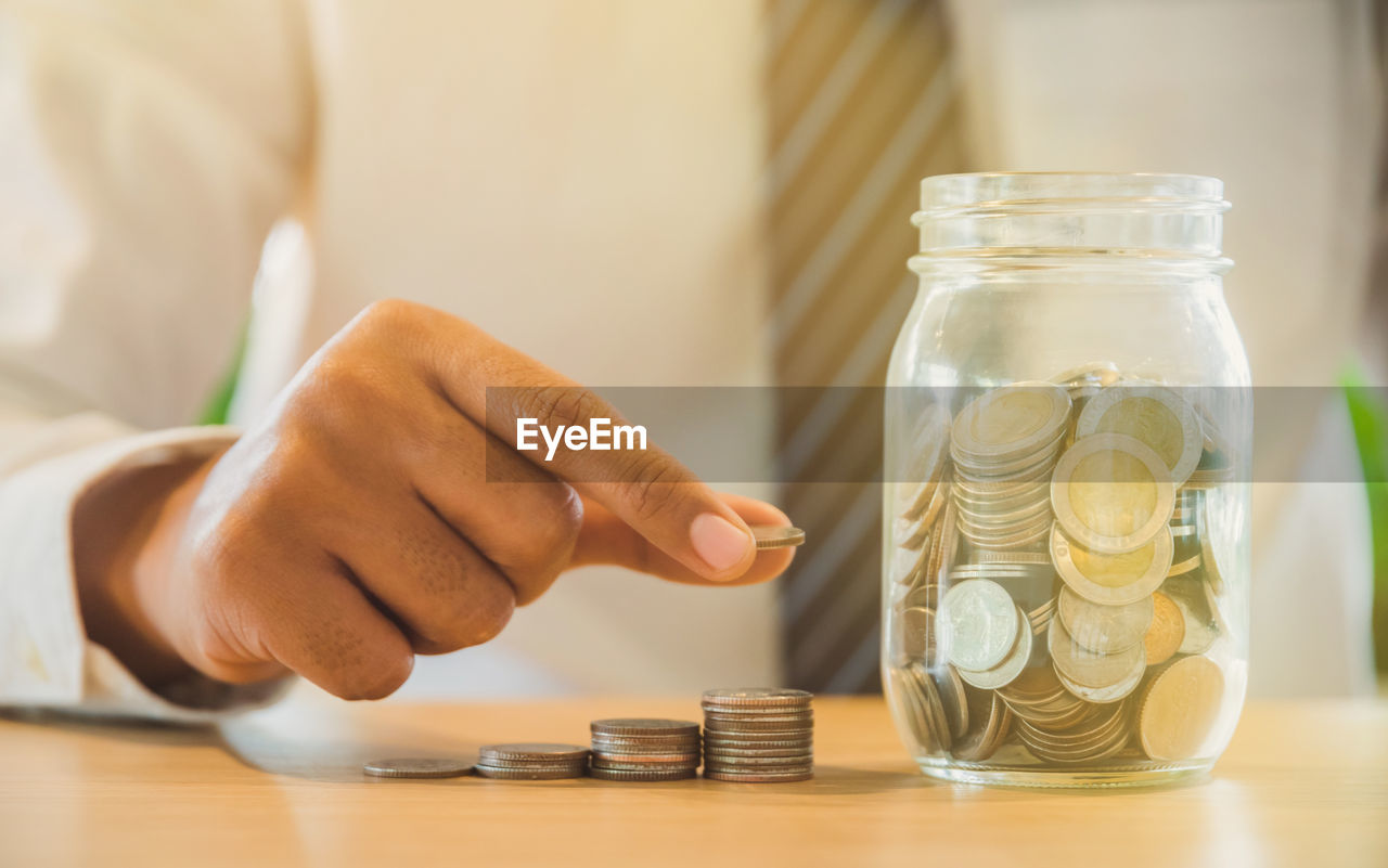 Midsection of man stacking coin by currency jar on table