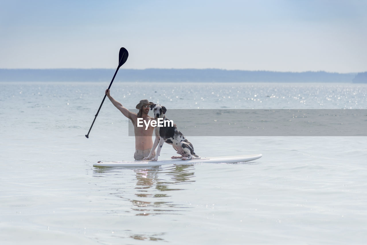 Shirtless man with dog paddleboarding in sea against sky