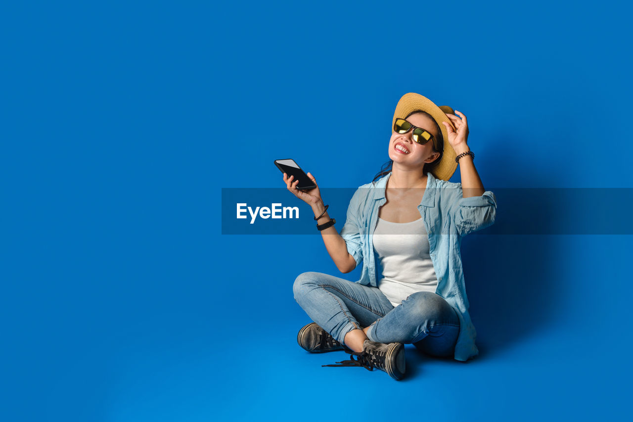 FULL LENGTH OF YOUNG WOMAN USING SMART PHONE WHILE SITTING ON BLUE WALL