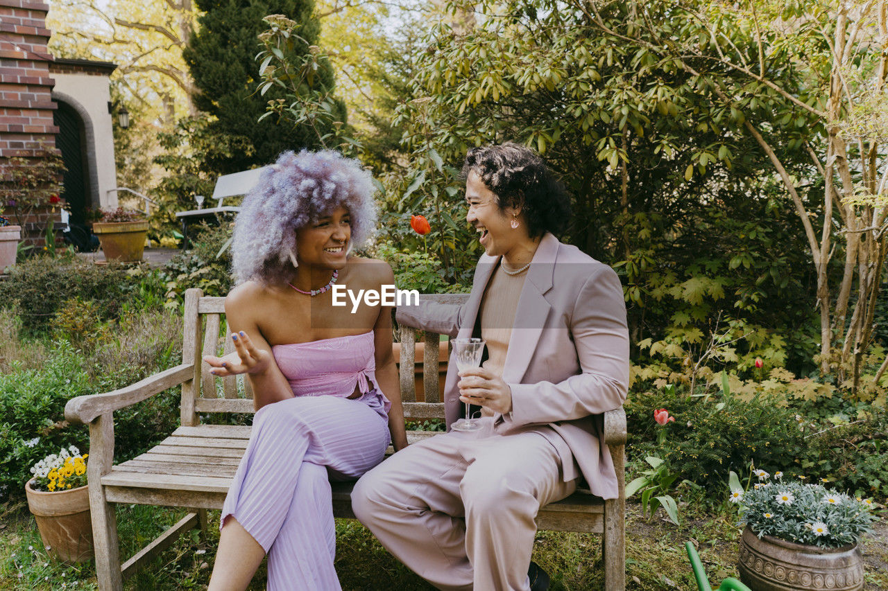 Smiling transwoman talking with happy gay friend while sitting on bench during dinner party in back yard