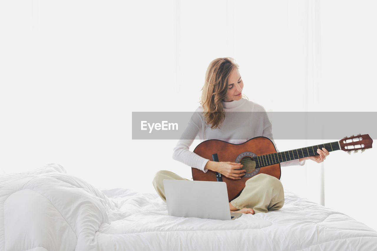 Woman playing guitar while sitting by laptop on bed