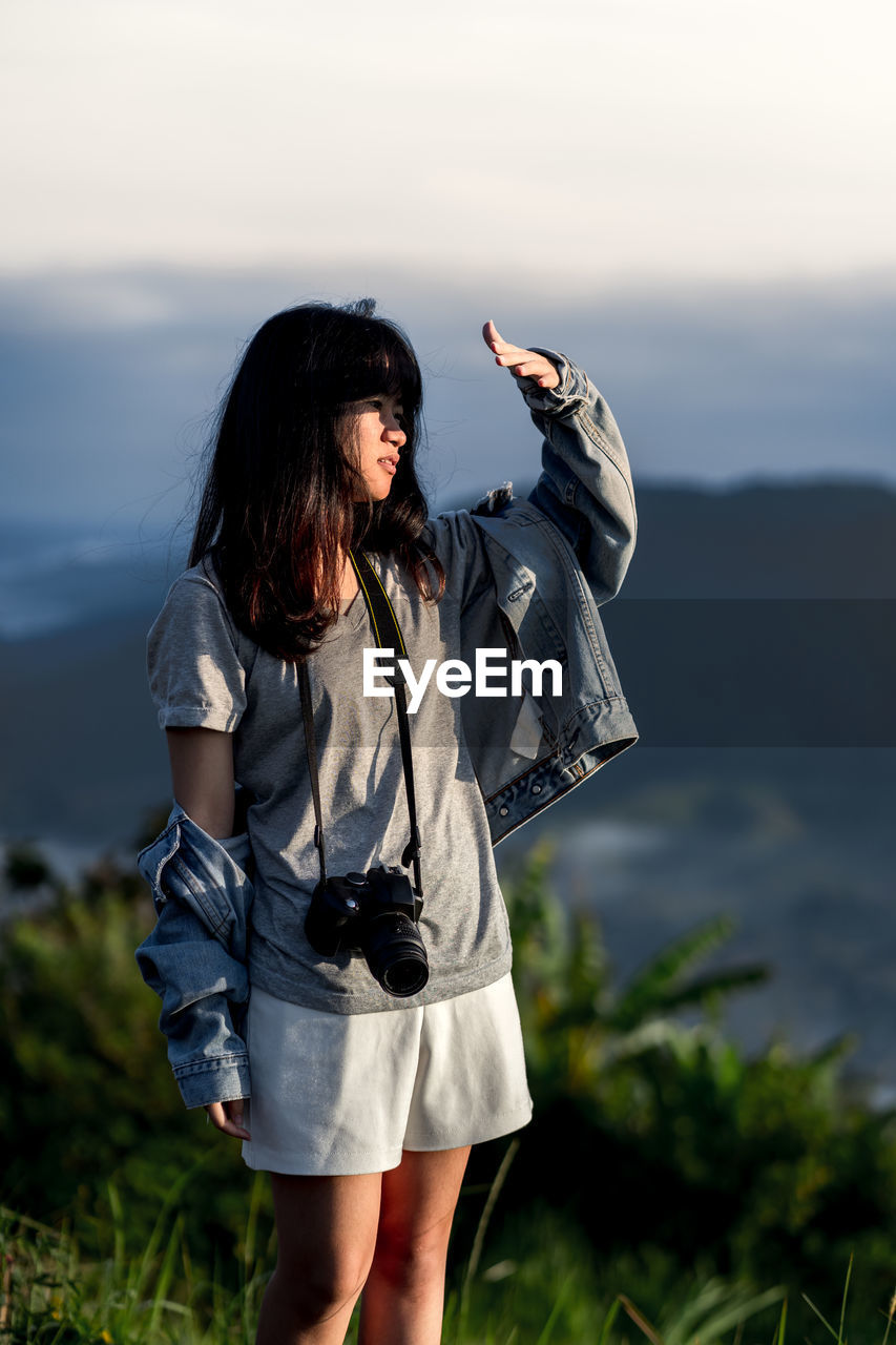 Young woman with digital camera standing against sky