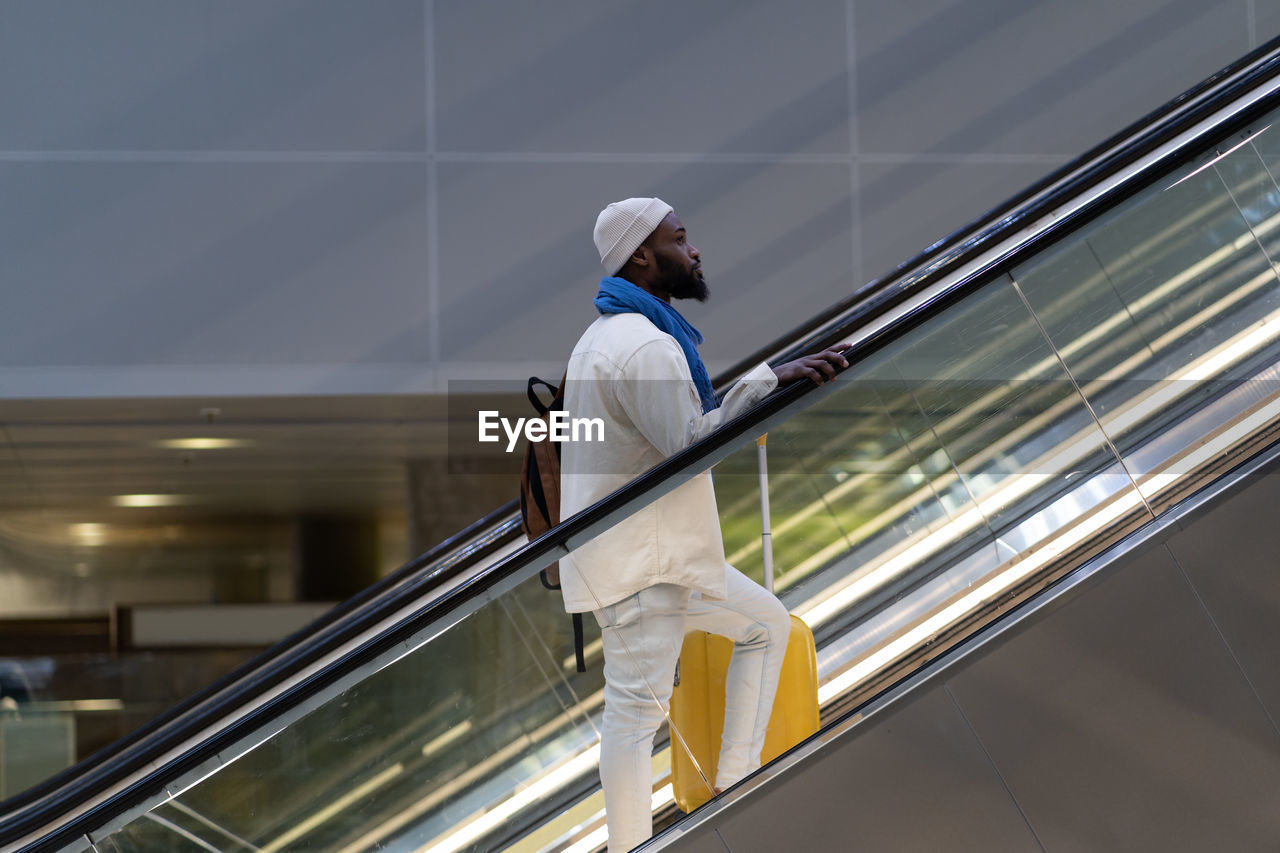African american passenger man with suitcase stands on escalator, holds handrail in airport terminal