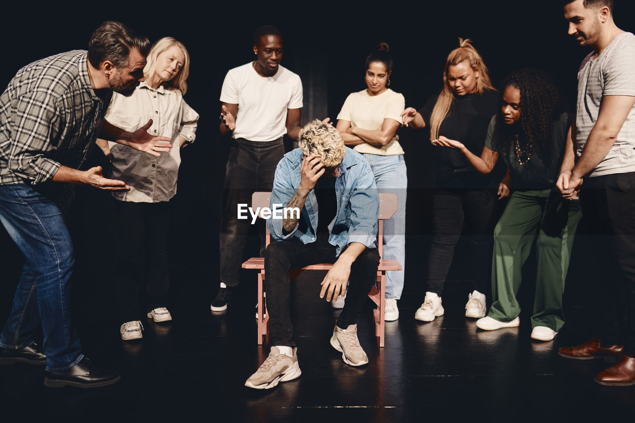 Distraught man scratching head while sitting amidst angry multiracial actors on stage