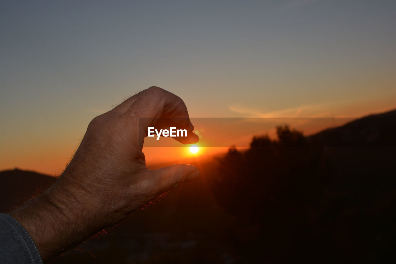 CLOSE-UP OF SILHOUETTE HAND HOLDING SUN AT SUNSET