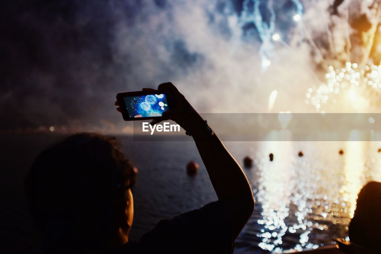 Rear view of person photographing through smart phone at fireworks