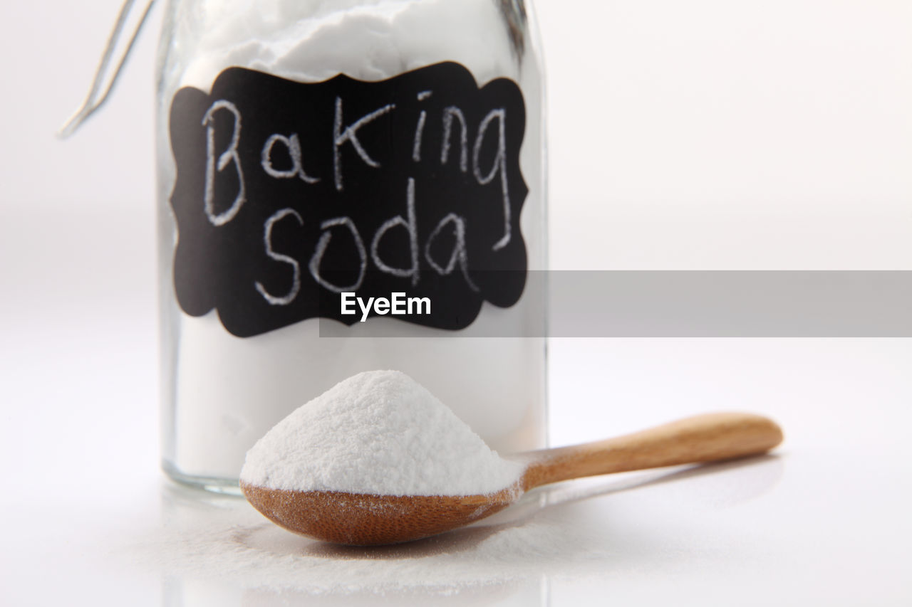 Close-up of baking soda in jar and wooden spoon against white background