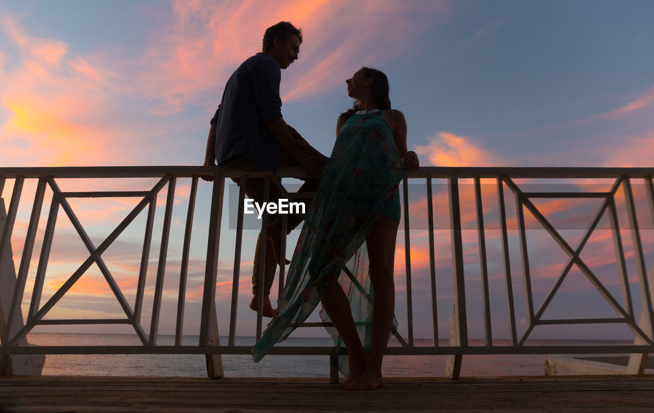 Low angle view of couple talking in balcony against cloudy sky during sunset