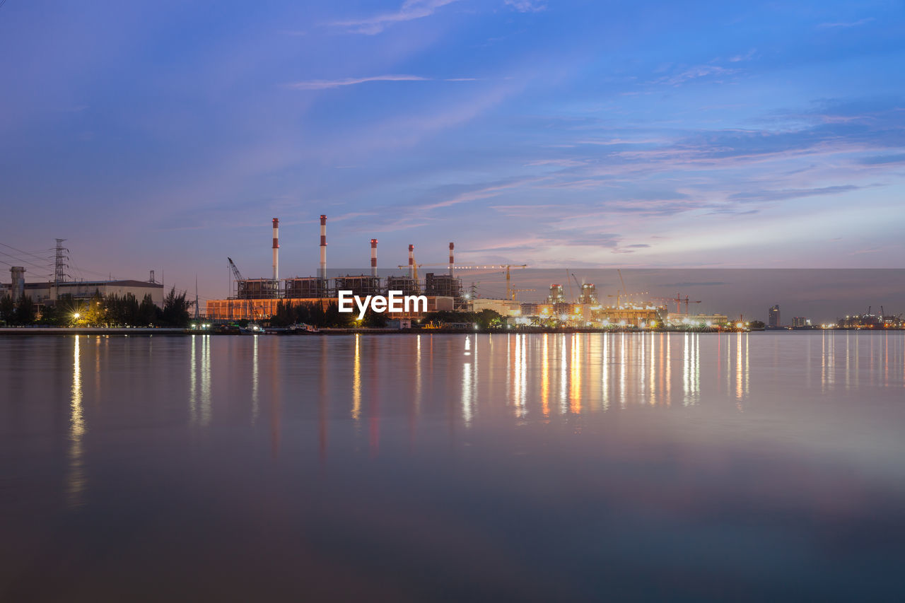 ILLUMINATED FACTORY BY SEA AGAINST SKY AT SUNSET