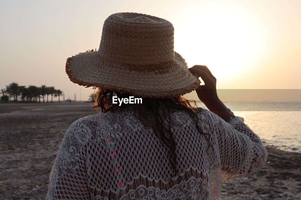 Rear view of woman wearing hat at beach against sky during sunset