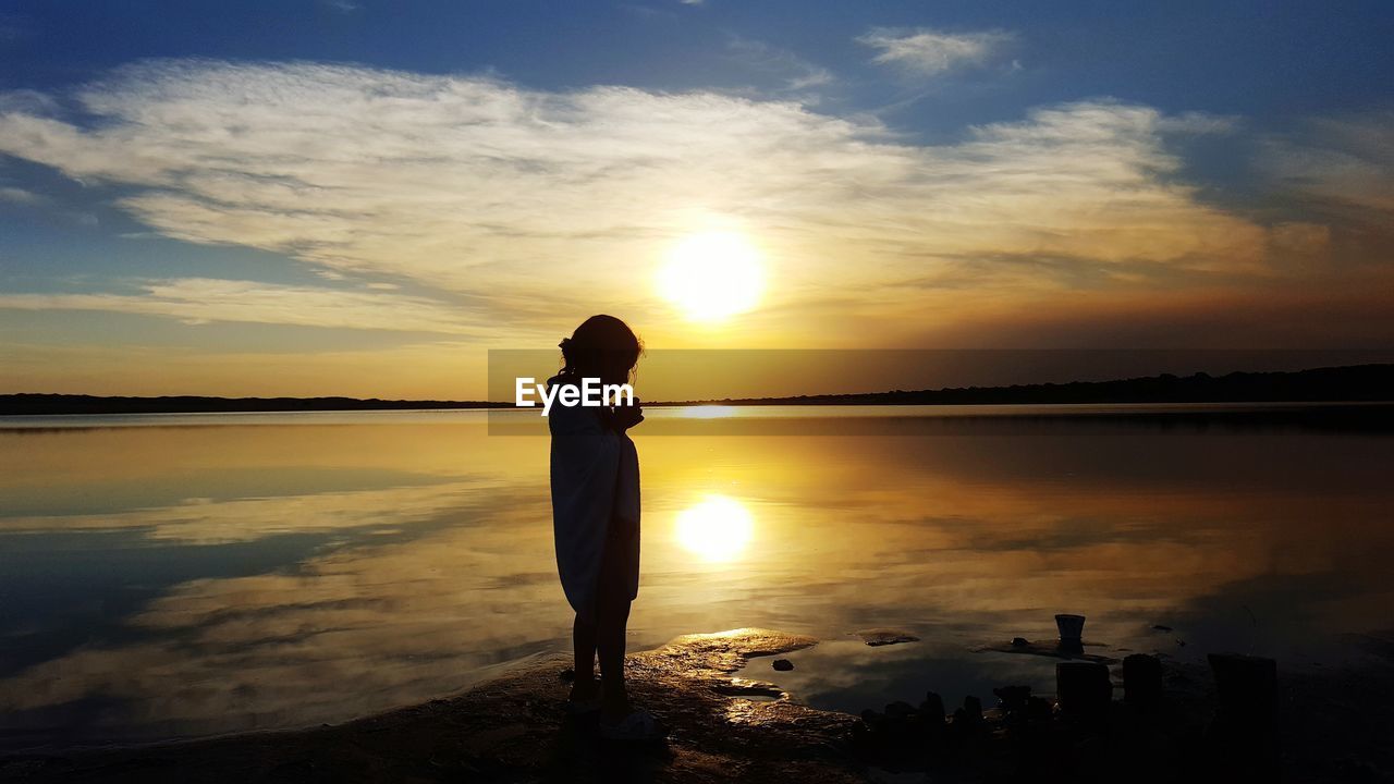 Silhouette of a girl standing on shore against sky during sunset