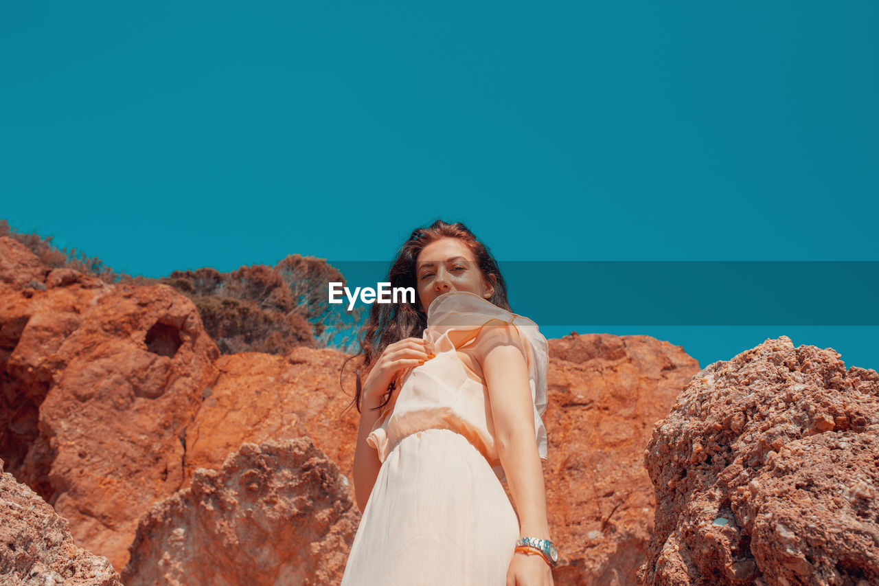 Low angle portrait of woman standing on rock against clear blue sky