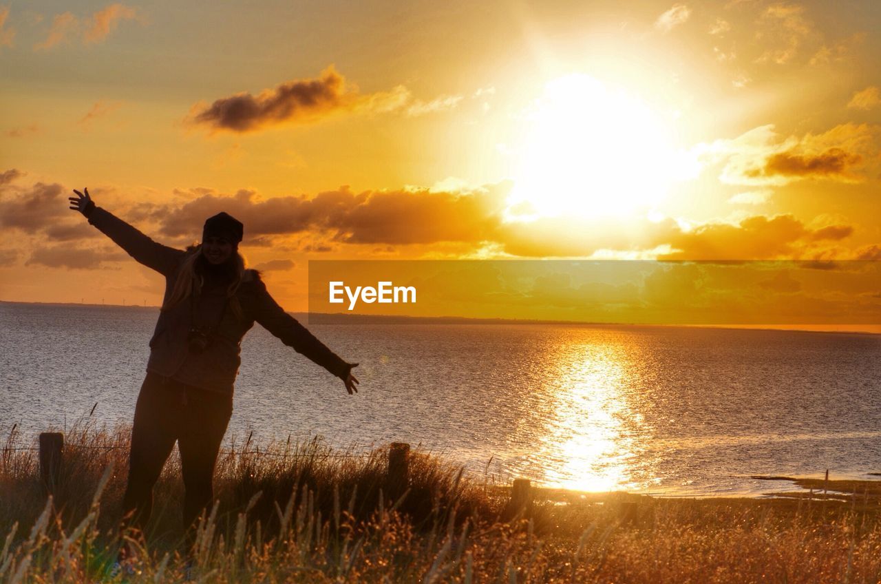 Woman standing on field by lake against sky during sunrise