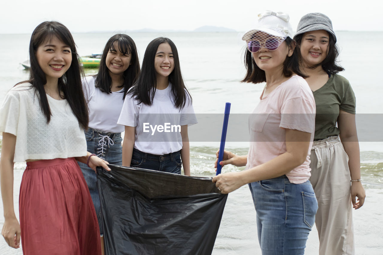 Portrait of smiling friends holding garbage bag while standing at beach against sky