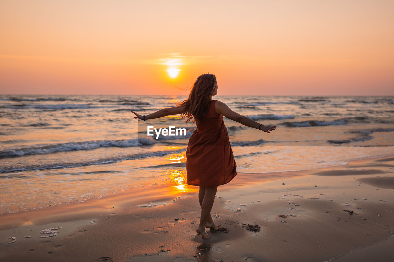 Rear view full length of woman with arms outstretched standing at beach during sunset
