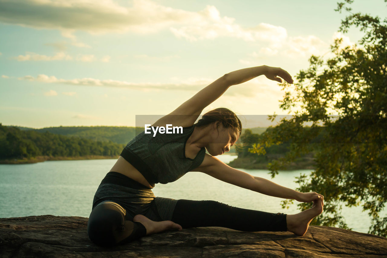 Young woman doing yoga by lake against sky